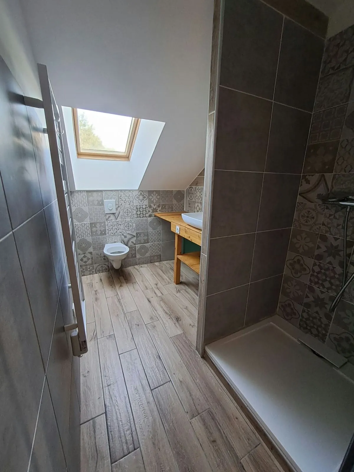 Salle de bain sous les combles, carreaux à motifs, douche, sol imitation bois et fenêtre de toit